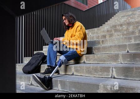 Disabled young man with leg prosthesis using laptop while sitting on steps Stock Photo