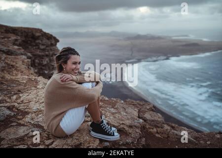 Smiling woman hugging knees while sitting on mountain against Famara Beach, Lanzarote, Spain Stock Photo