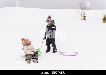 Brother and sister playing with sled on snow covered landscape Stock Photo