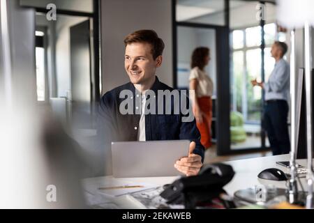 Smiling businessman looking away while sitting with colleagues in background at open plan office Stock Photo
