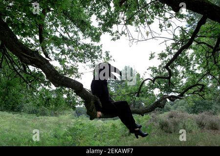 Woman in crow costume sitting on branch of tree in forest against sky Stock Photo