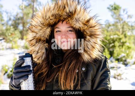 Smiling woman wearing fur hooded winter coat holding bottle while standing at forest Stock Photo