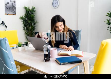 Smiling student with laptop reading book while sitting on table against white wall at home Stock Photo