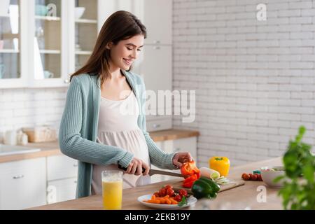 smiling pregnant woman cutting fresh vegetables in kitchen Stock Photo