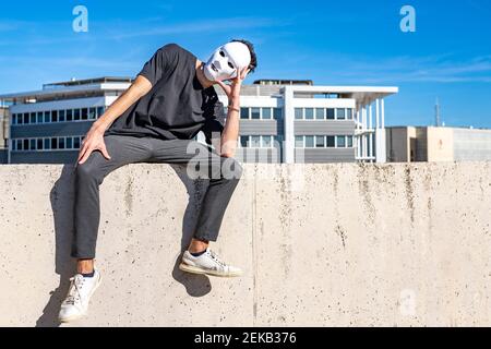 Man wearing white mask looking away while sitting with head in hands on wall Stock Photo