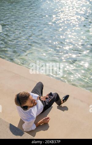 Businessman with mobile phone and headphones looking away while sitting on steps by lake Stock Photo