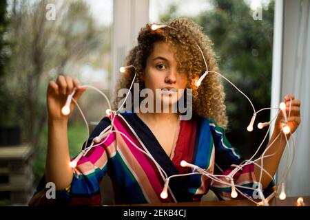 Confused curly haired woman holding illuminated Christmas lights at home Stock Photo