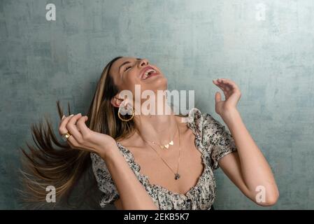Cheerful woman tossing hair while sitting against concrete wall Stock Photo