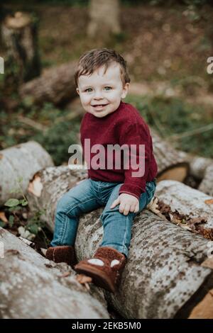 Confused toddler boy staring while sitting on log in forest during autumn Stock Photo