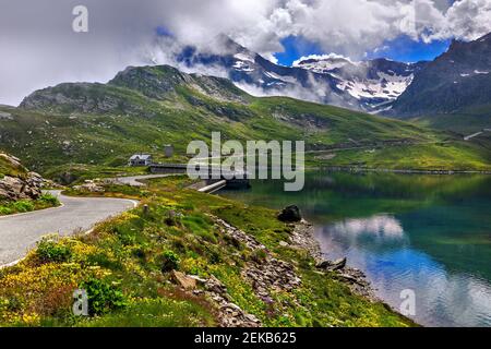 View of road along green shores and small alpine lake as mountains on background in Piedmont, Northern Italy. Stock Photo