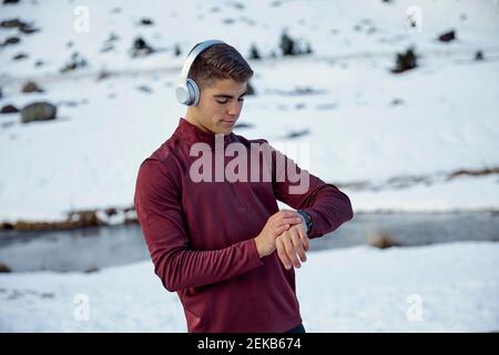 Young male athlete checking time in smartwatch while wearing headphones during winter Stock Photo