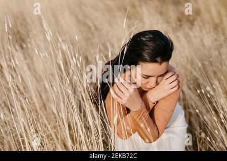 Thoughtful young woman sitting amidst grass in field at sunset Stock Photo