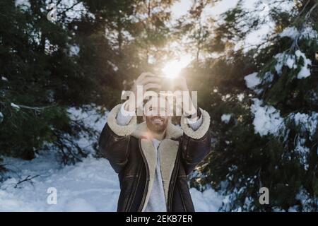 Man wearing jacket photographing while standing against trees during winter Stock Photo