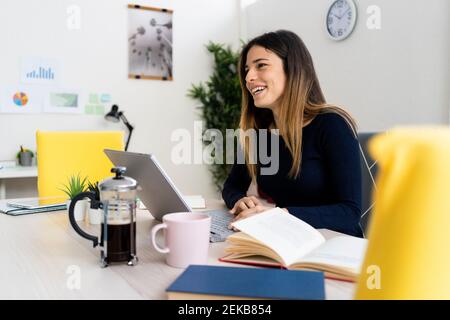 Happy young female freelance worker with laptop and book looking away at home Stock Photo