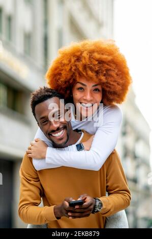 Smiling woman piggybacking on man using mobile phone while standing outdoors Stock Photo