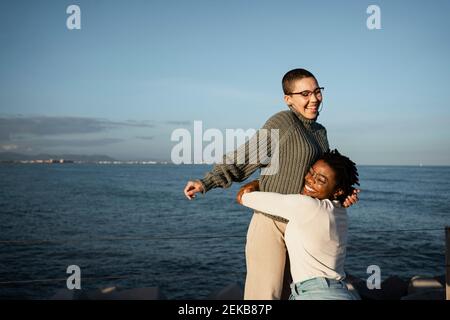 Smiling woman embracing friend standing against sea Stock Photo