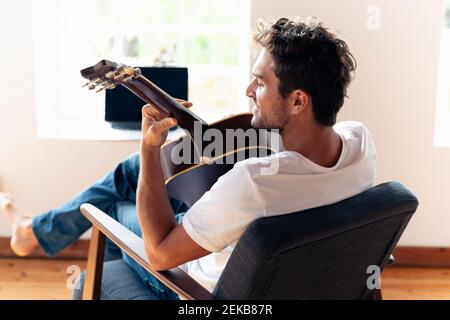 Smiling man playing guitar while relaxing on armchair at home Stock Photo