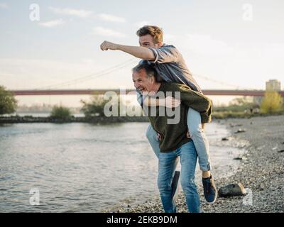 Happy father giving piggyback ride to son at riverbank against sky Stock Photo