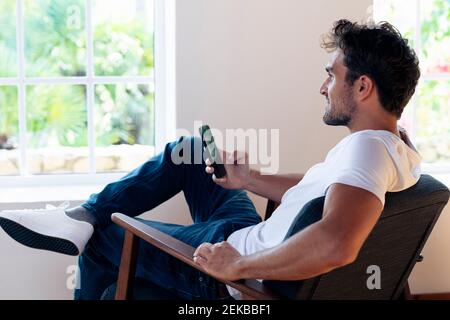 Man with smart phone looking through window while relaxing on armchair at home Stock Photo