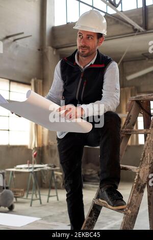 Male engineer holding blueprint looking away while standing on ladder in building Stock Photo
