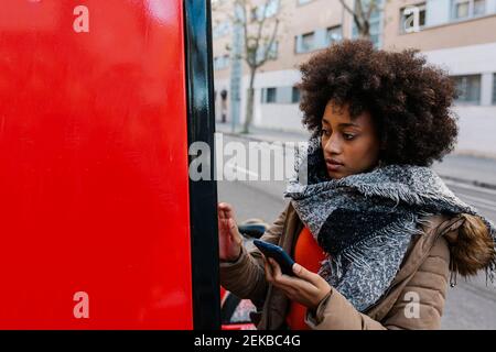 Afro woman with smart phone buying ticket from machine during winter Stock Photo