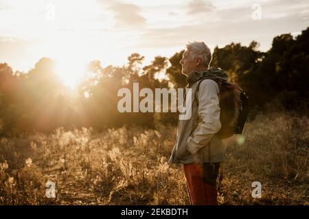 Senior male hiker with hands in pockets standing on agricultural field during sunset Stock Photo