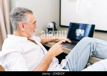 Mature man using laptop with feet up at desk while having coffee in hotel room Stock Photo