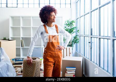 Smiling Afro woman looking with carpet looking through window in new loft apartment Stock Photo