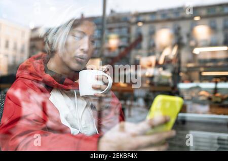 Young man using smart phone while having coffee seen through glass in cafe Stock Photo
