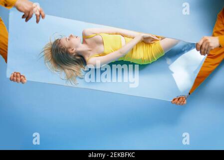 Young woman lying on blue background while reflected in mirror being held by men Stock Photo