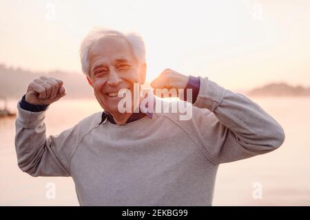 Smiling senior man showing bicep while standing against sky during sunrise Stock Photo