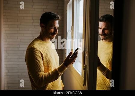 Serious man using smart phone while standing by window at home Stock Photo