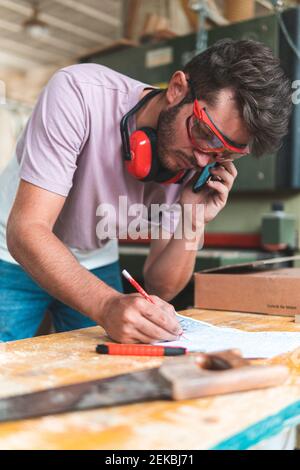 Young male craftsperson talking on smart phone while writing on paper in workshop Stock Photo