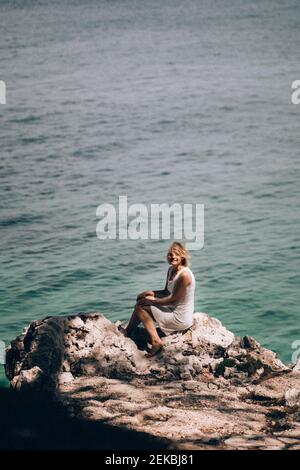 Mature woman sitting on rock at beach against seascape during sunny day Stock Photo