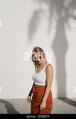 Smiling beautiful woman looking down against tree shadow on white wall Stock Photo