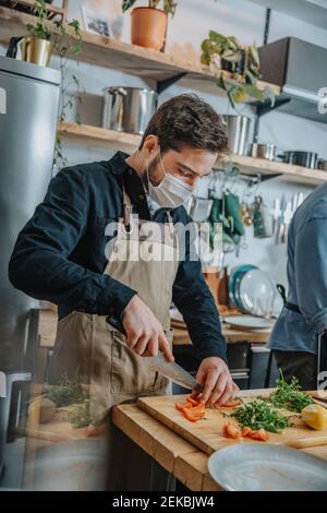 Mid adult chef wearing protective face mask cutting vegetable while standing with colleague in kitchen Stock Photo