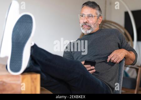 Thoughtful businessman using mobile phone while sitting with legs on table at home Stock Photo