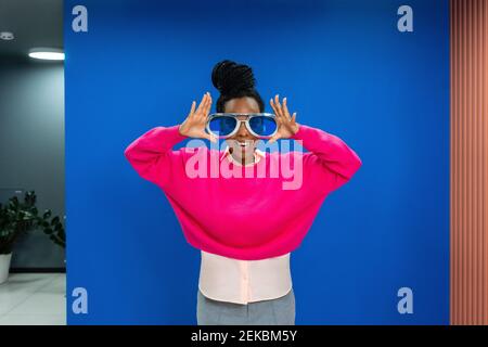 Cheerful creative female professional wearing oversized sunglasses against blue wall at work place Stock Photo
