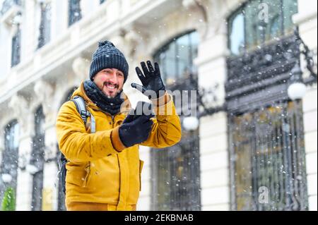 Smiling man waving hand to mobile phone while talking on video call by building during winter Stock Photo