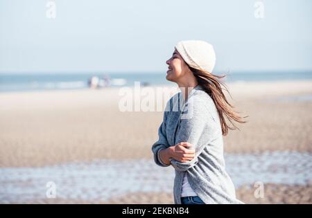 Happy young woman in knit hat hugging self while standing at beach Stock Photo