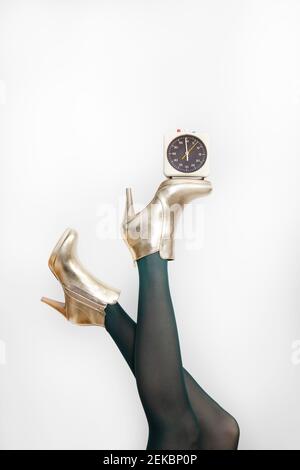 Woman wearing golden stilettos while balancing alarm clock against white background Stock Photo