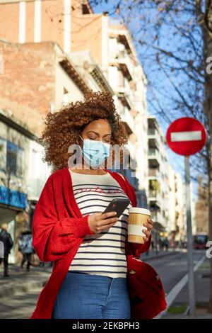 Young woman wearing face mask using mobile phone while standing with coffee cup in city Stock Photo