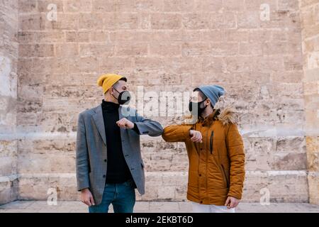 Young men wearing knit hat and protective face mask giving elbow bump while greeting against wall Stock Photo