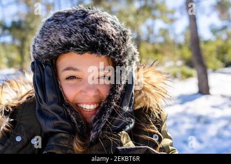 Cheerful woman wearing fur cap smiling while standing on forest during winter Stock Photo