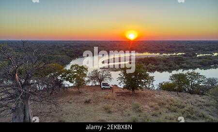 Aerial view of a jeep with a rooftop tent close to the river and a big baobab tree at sunset, Cunene river area, Angola Stock Photo