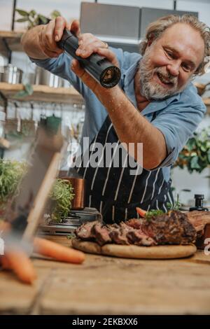 Smiling chef seasoning pepper on grilled tomahawk steak while standing in kitchen Stock Photo
