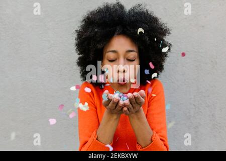 Afro young woman with eyes closed blowing confetti against wall Stock Photo