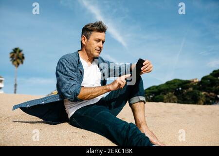 Mature man using mobile phone while sitting at beach during sunny day Stock Photo