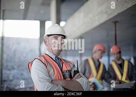 Confident male contractor with arms crossed at site with construction workers seen in background Stock Photo