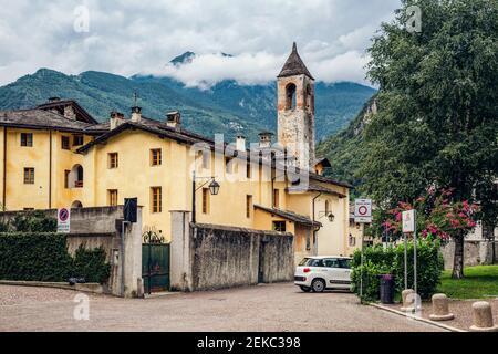 Bell tower by tree in town against mountain range, Valchiavenna, Chiavenna, Province of Sondrio, Lombardy, Italy Stock Photo